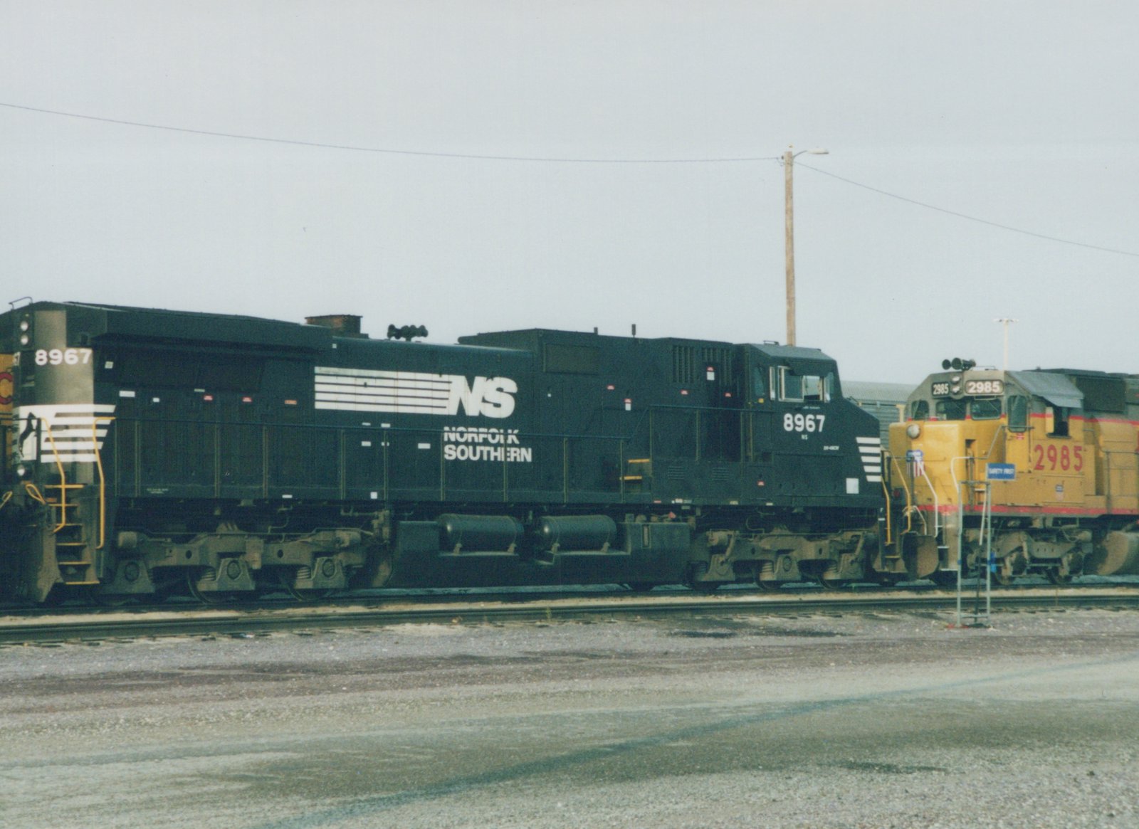 Side view of a black Norfolk Southern locomotive numbered 8967 in a railyard, with a yellow locomotive numbered 2985 on an adjacent track under an overcast sky.