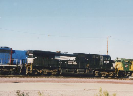 A black Norfolk Southern locomotive numbered 8986 sits on tracks in a rail yard under a clear sky, with green and yellow and blue locomotives visible behind it.