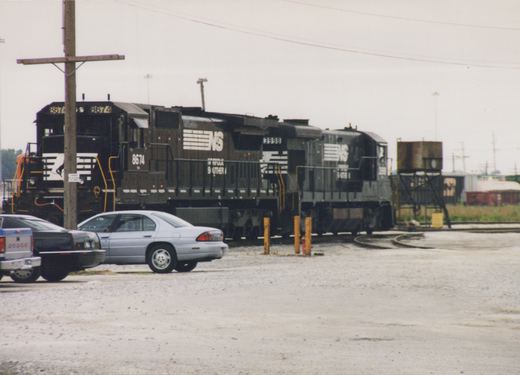 A three-quarter view of two black Norfolk Southern GP38-2 diesel locomotives, numbered 8674 and 3998, in a gravel rail yard with a light blue sedan and a white pickup truck parked in front.