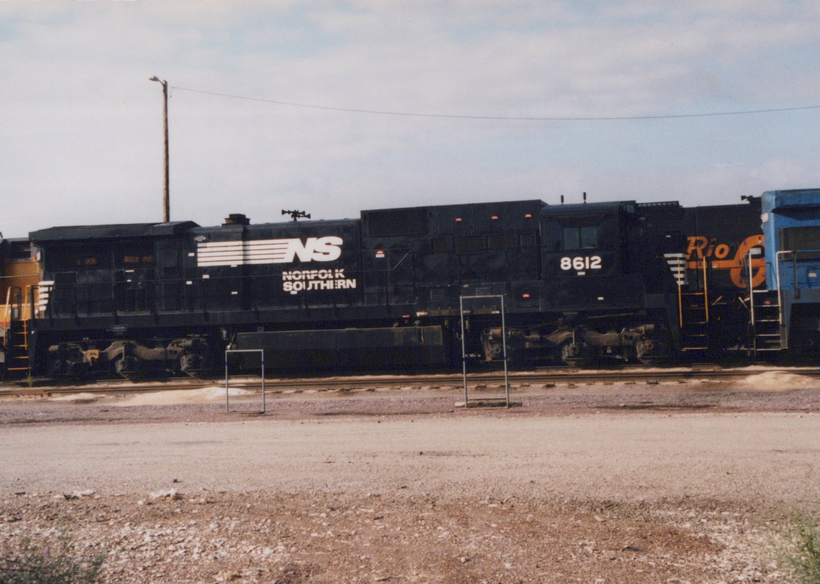 A black Norfolk Southern diesel locomotive, number 8612, is parked in a rail yard. Another locomotive with Rio Grande lettering is partially visible behind it.