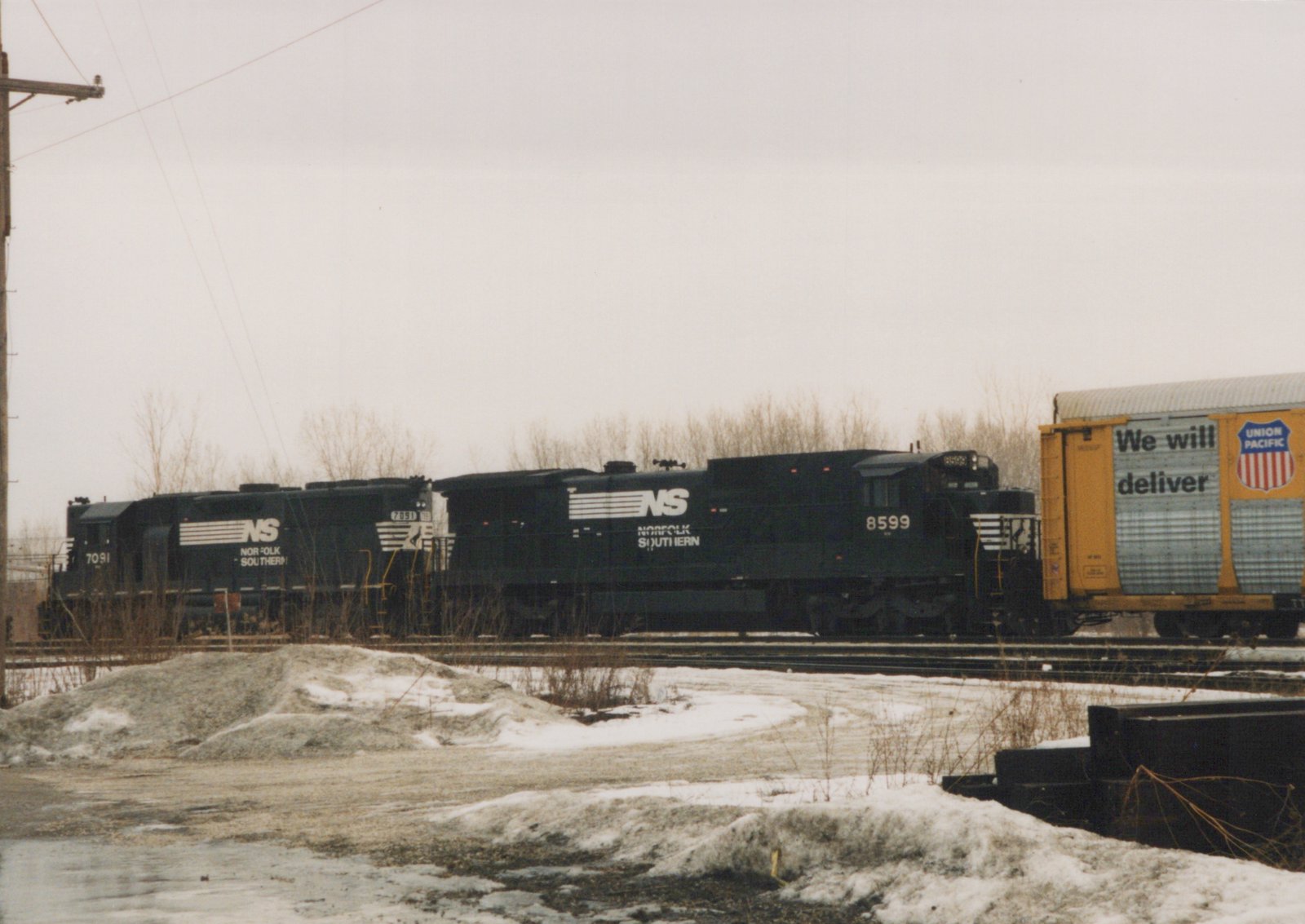 Two black Norfolk Southern locomotives with numbers 8599 and 7091 are pulling a yellow Union Pacific freight car on a snowy railroad yard.