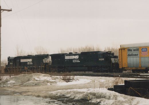 Two black Norfolk Southern locomotives with numbers 8599 and 7091 are pulling a yellow Union Pacific freight car on a snowy railroad yard.