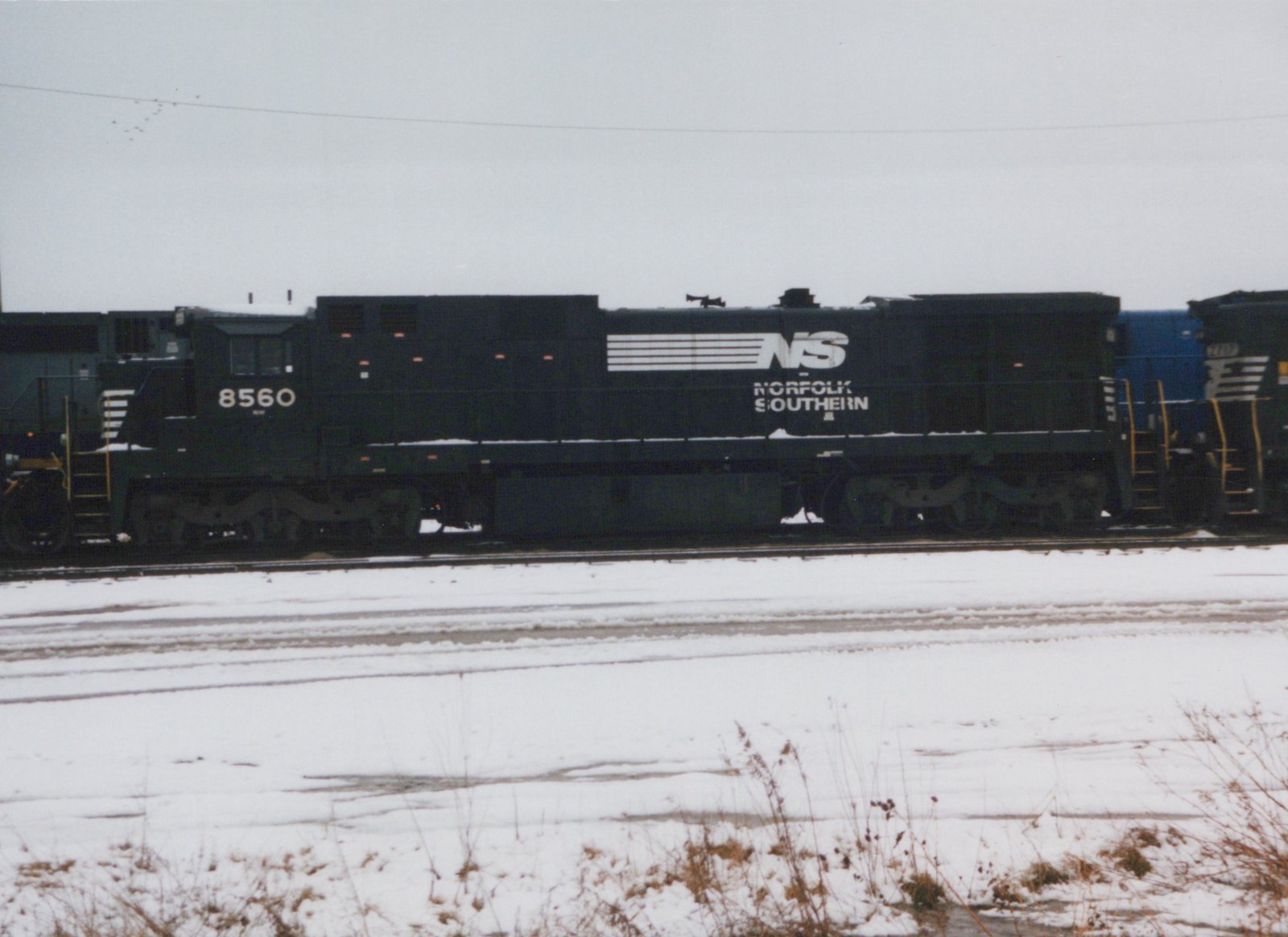 Side view of a dark green Norfolk Southern locomotive numbered 8560, sitting on tracks in a snow-covered yard under an overcast sky.