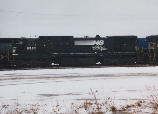 Side view of a dark green Norfolk Southern locomotive numbered 8560, sitting on tracks in a snow-covered yard under an overcast sky.