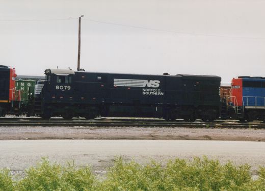 A black Norfolk Southern diesel locomotive numbered 8079 sits on tracks in a railyard next to another locomotive and freight cars under an overcast sky.