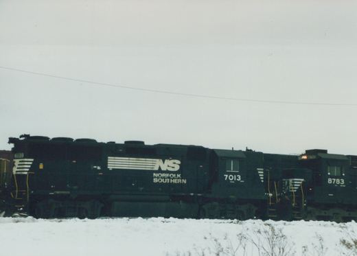 Two black Norfolk Southern locomotives numbered 7013 and 8783 are parked side by side on a snow-covered railroad yard under an overcast sky.