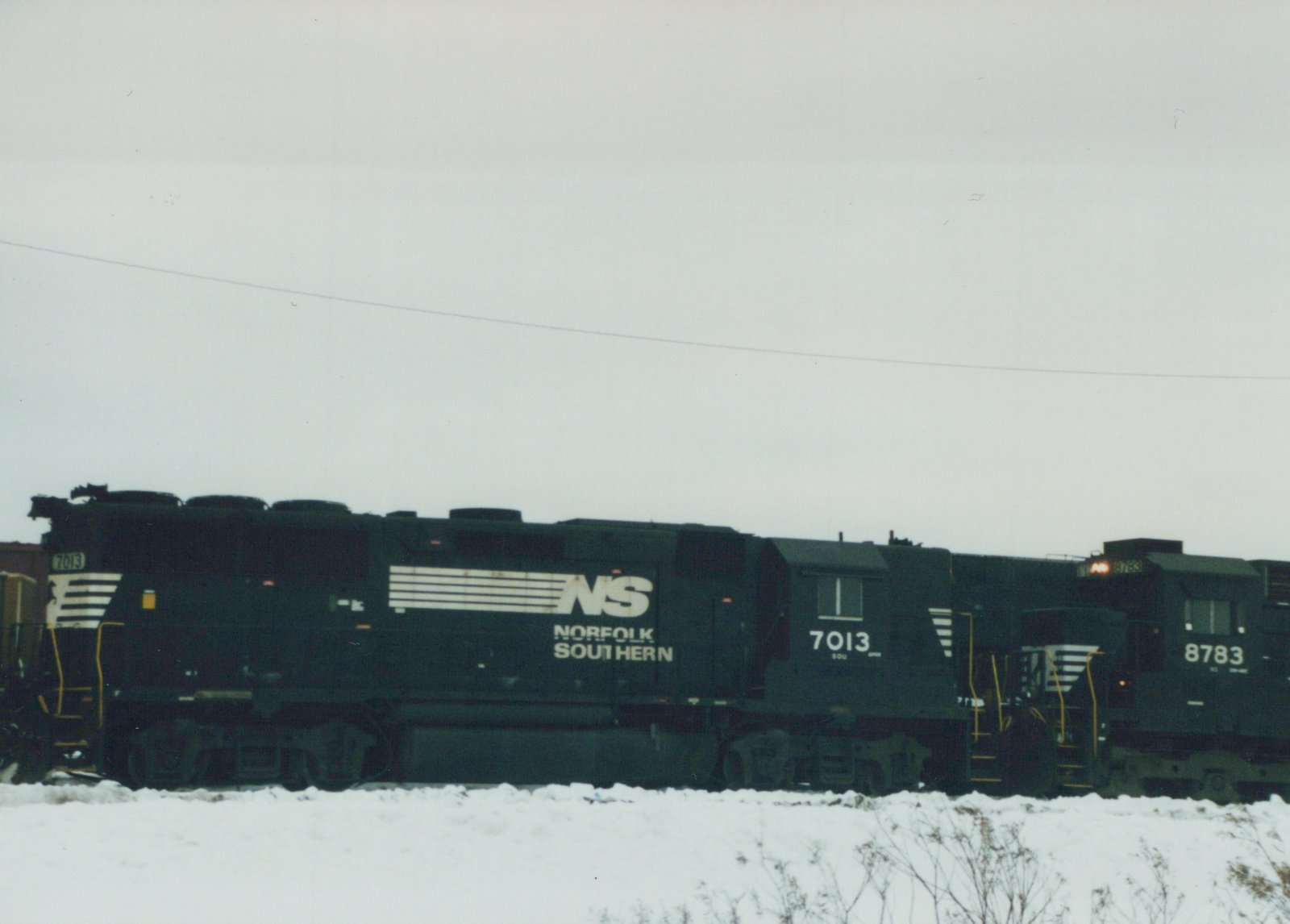 Two black Norfolk Southern locomotives numbered 7013 and 8783 are parked side by side on a snow-covered railroad yard under an overcast sky.