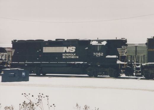 A black Norfolk Southern locomotive numbered 7062 is parked in a snow-covered yard, with the company's white 'NS' logo and lettering visible on its side.