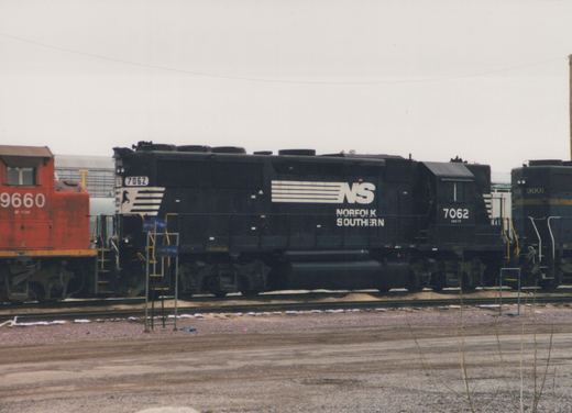 A black Norfolk Southern diesel locomotive with the number 7062 is parked on tracks in a yard alongside other locomotives, with patches of snow on the ground.