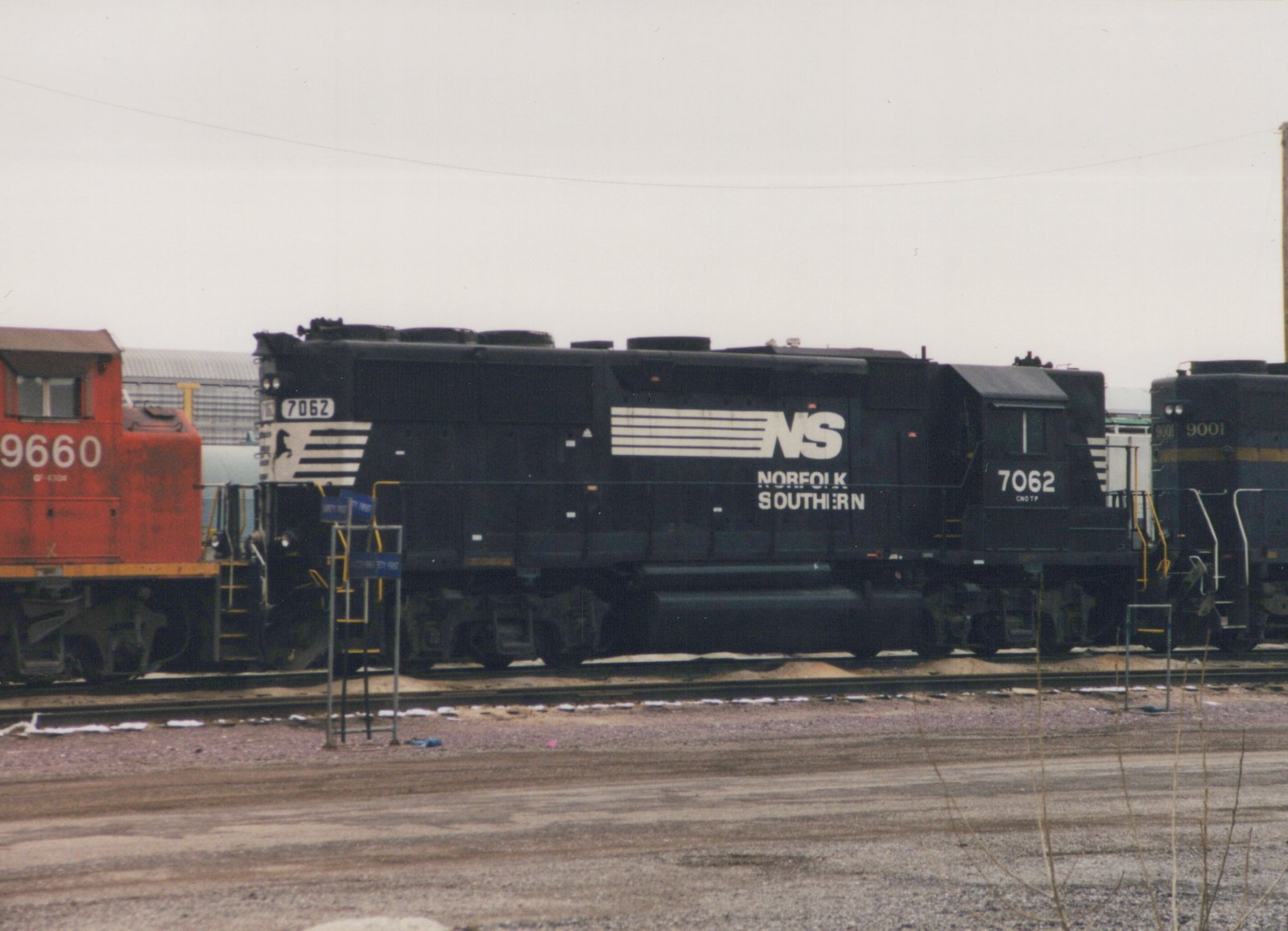 A black Norfolk Southern diesel locomotive with the number 7062 is parked on tracks in a yard alongside other locomotives, with patches of snow on the ground.