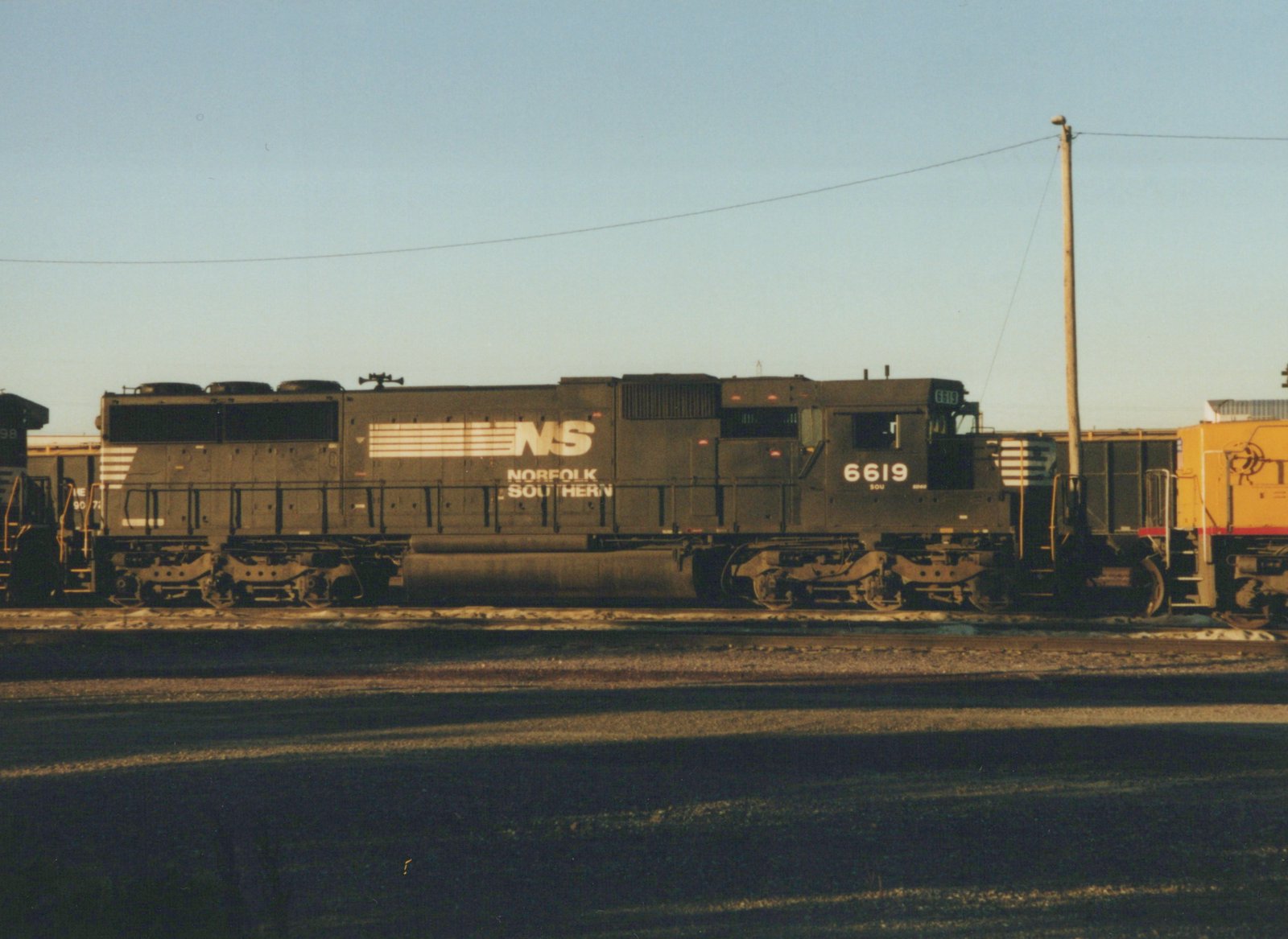 A side view of a dark gray Norfolk Southern locomotive number 6619, with another locomotive partially visible to the left and a yellow one to the right, in a yard under a clear sky.