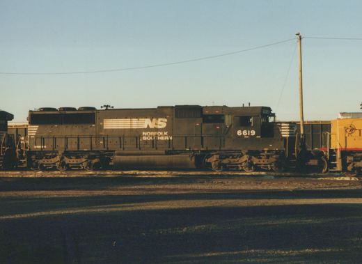 A side view of a dark gray Norfolk Southern locomotive number 6619, with another locomotive partially visible to the left and a yellow one to the right, in a yard under a clear sky.