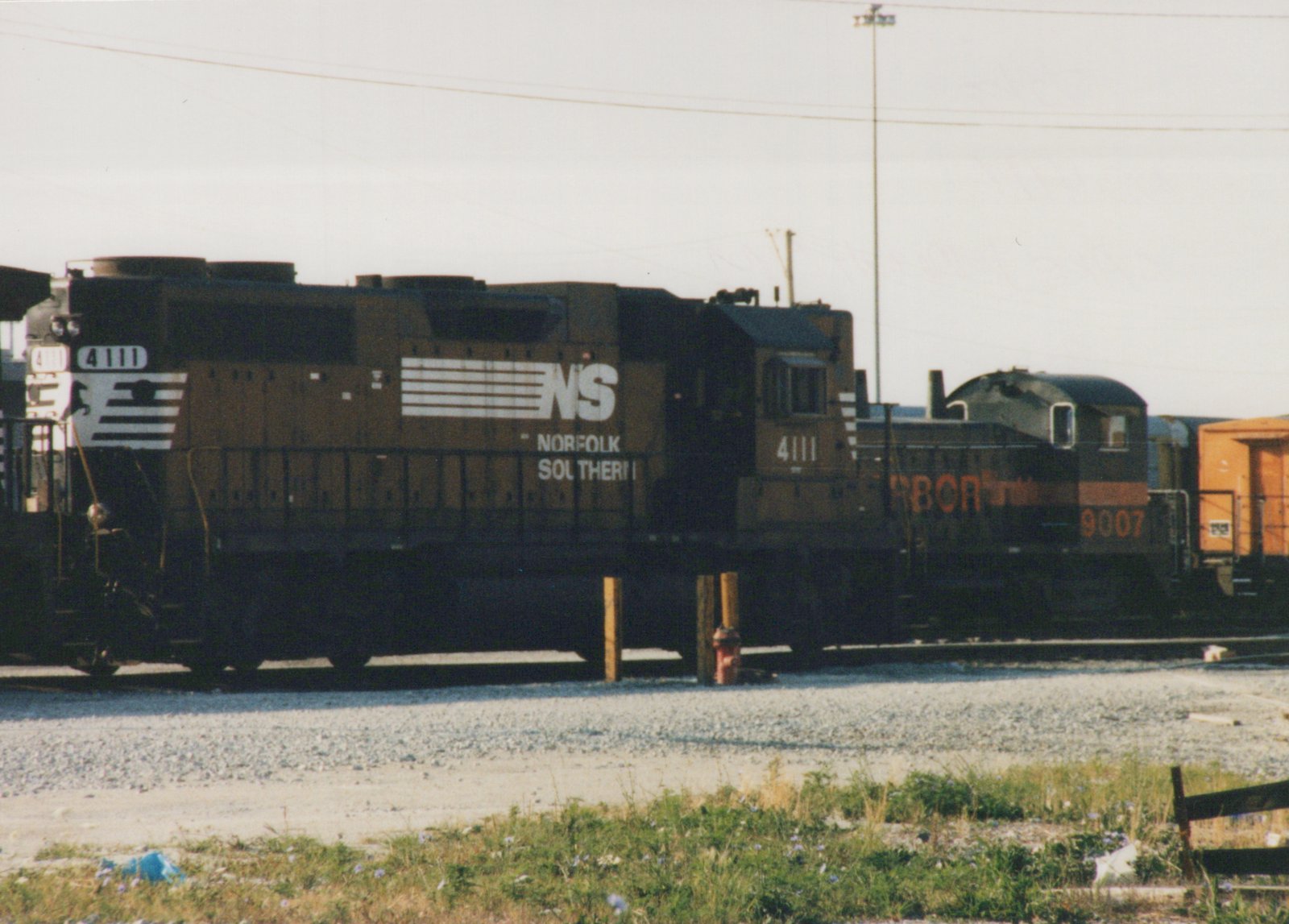 Two diesel locomotives, one Norfolk Southern numbered 4111 and another Rio Grande numbered 9007, parked on parallel tracks in a gravel yard under an overcast sky.
