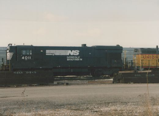 A dark blue Norfolk Southern diesel locomotive numbered 4011 is parked in a yard, with a yellow Union Pacific locomotive behind it, under an overcast sky.