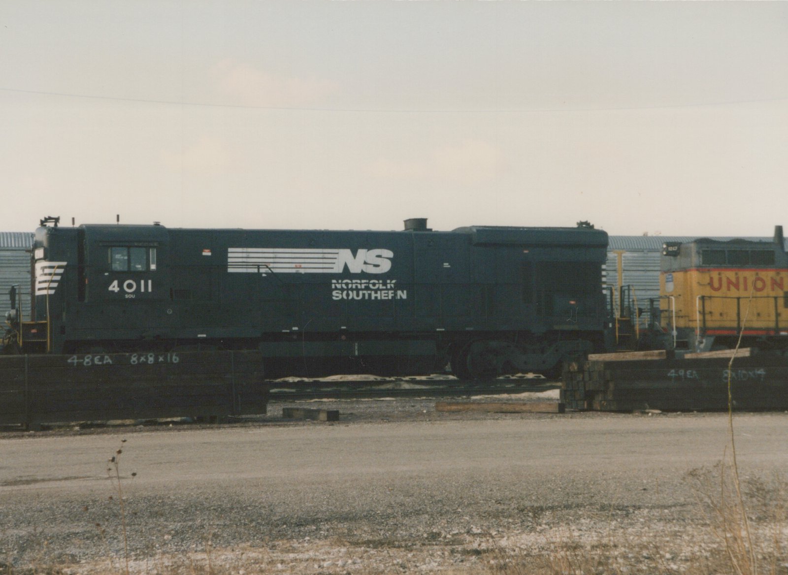 A dark blue Norfolk Southern diesel locomotive numbered 4011 is parked in a yard, with a yellow Union Pacific locomotive behind it, under an overcast sky.