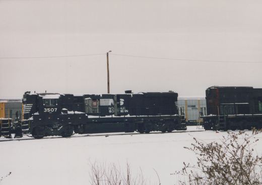 A side view of a black Norfolk Southern GP38-2 diesel locomotive, number 3507, in a snowy yard with freight cars and a gray, overcast sky.