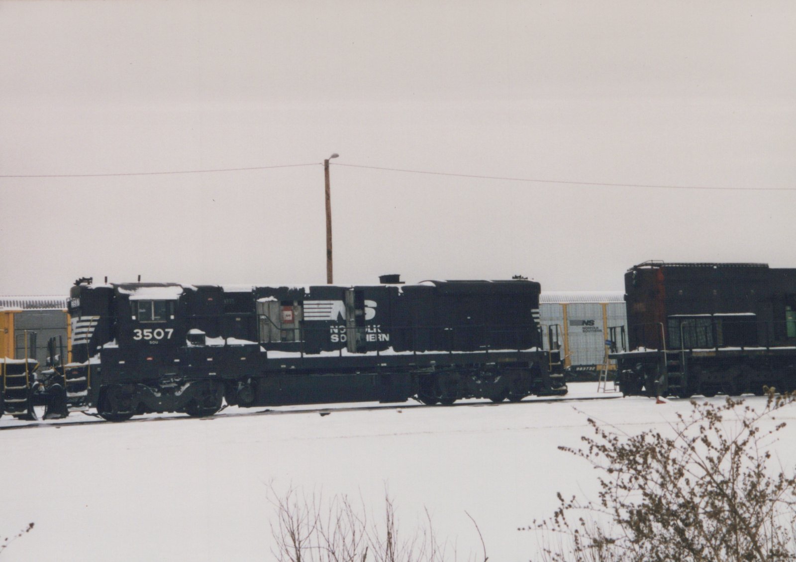 A side view of a black Norfolk Southern GP38-2 diesel locomotive, number 3507, in a snowy yard with freight cars and a gray, overcast sky.