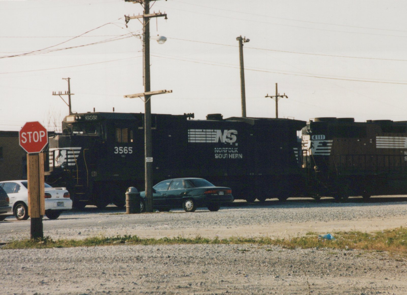 A black Norfolk Southern freight train with two locomotives passes a red stop sign next to parked cars on a gravel lot, under a clear sky.