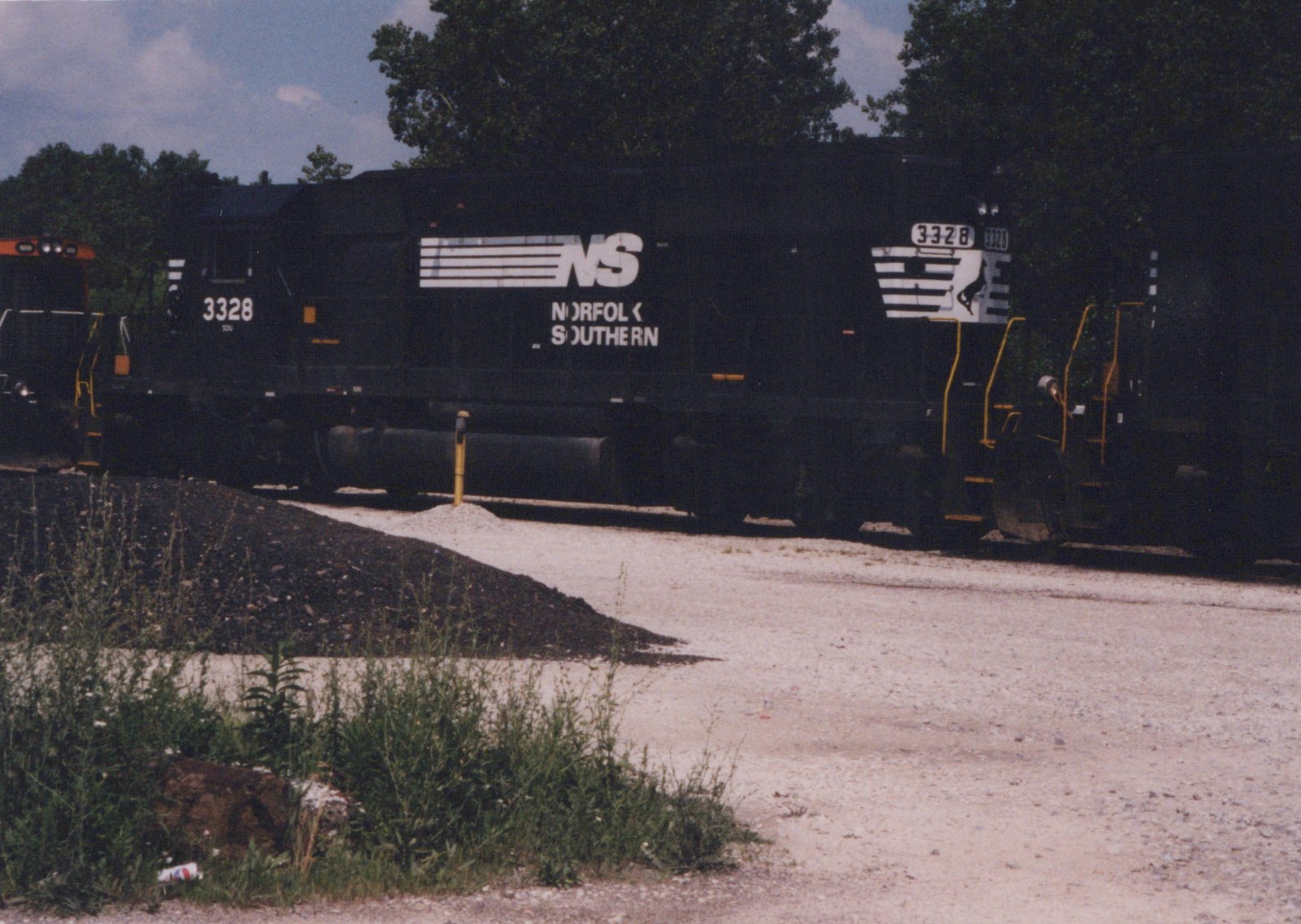A black Norfolk Southern locomotive with the number 3328, featuring white lettering and stripes, sits on a track in a gravel yard surrounded by trees and weeds, under a partly cloudy sky.