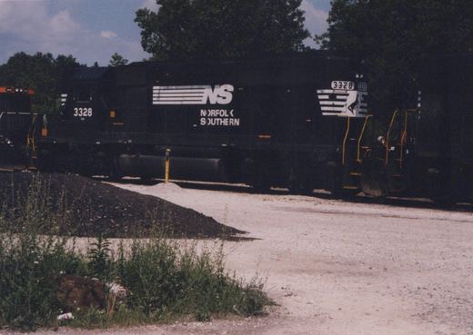 A black Norfolk Southern locomotive with the number 3328, featuring white lettering and stripes, sits on a track in a gravel yard surrounded by trees and weeds, under a partly cloudy sky.