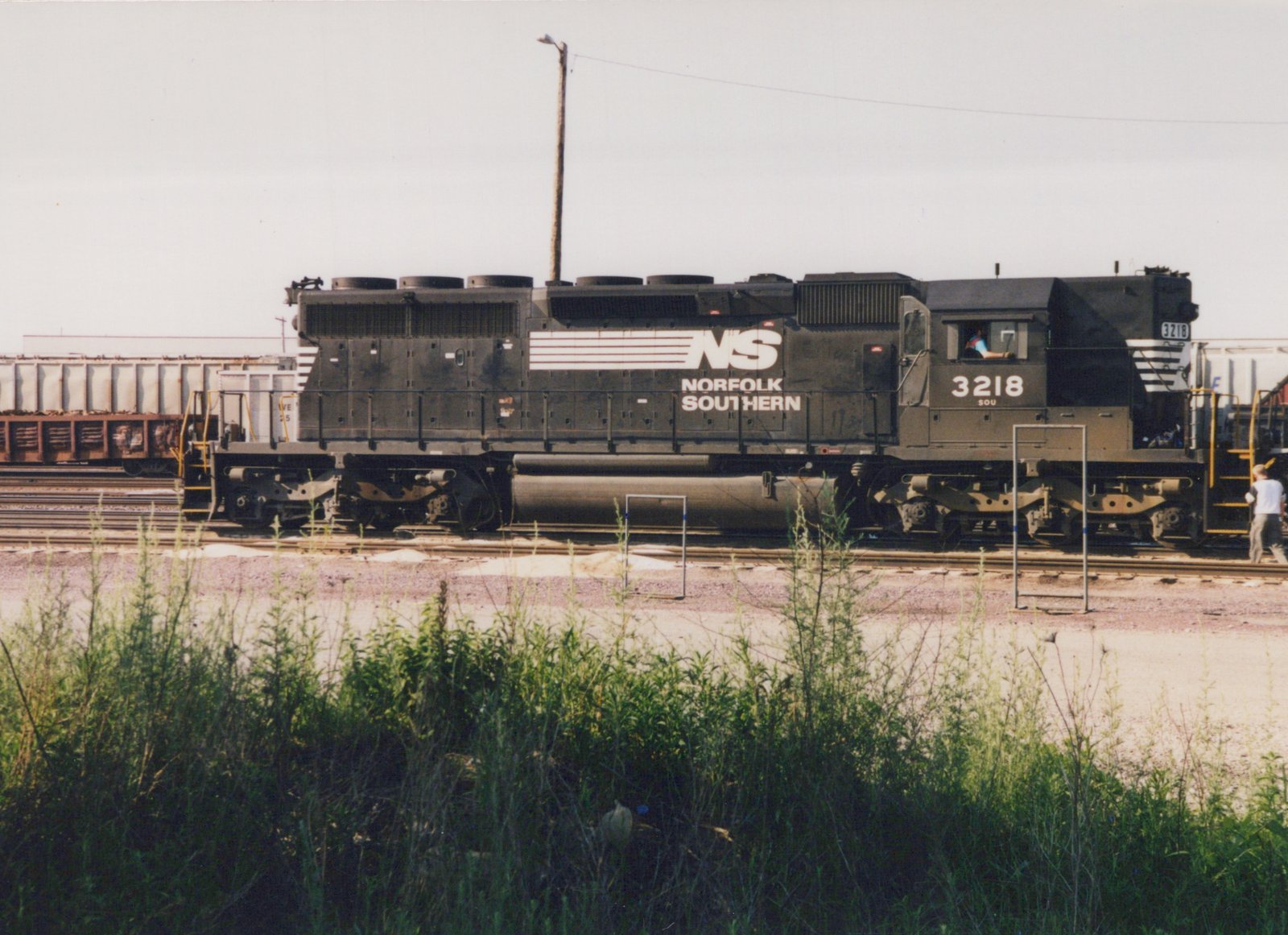 A black Norfolk Southern diesel locomotive numbered 3218 sits on tracks in a rail yard, surrounded by weeds, with a person walking beside it and another in the cab.