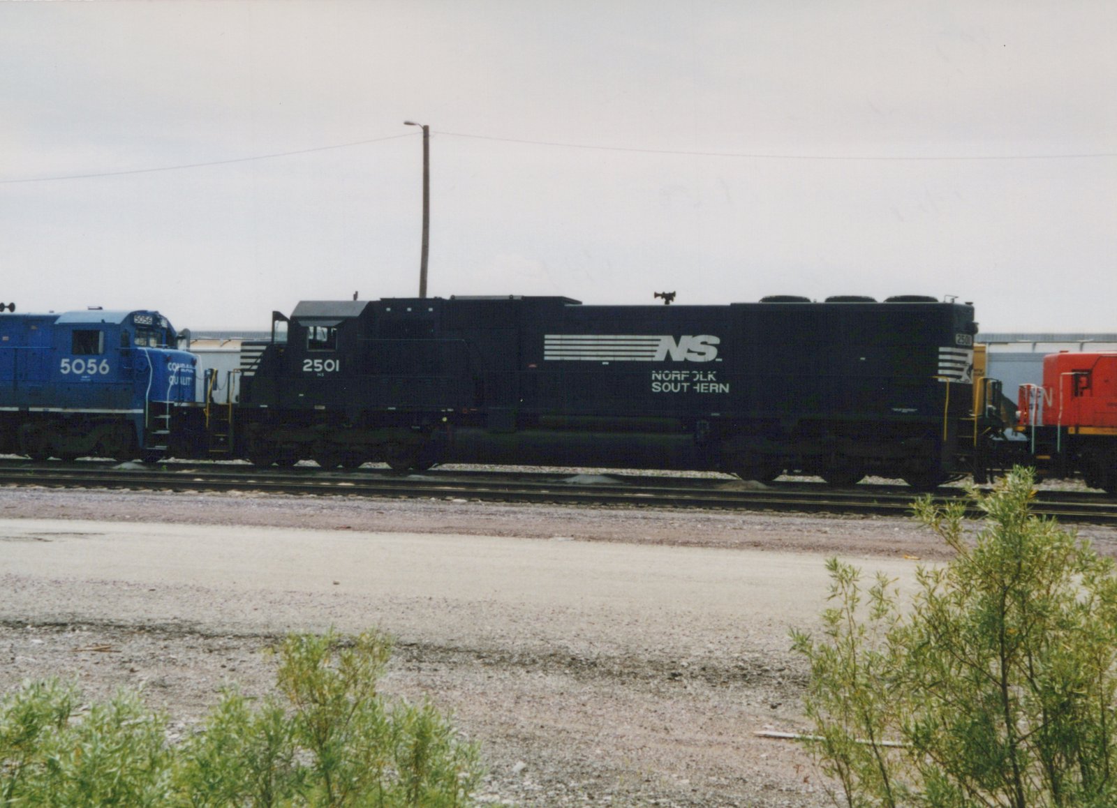 A black Norfolk Southern diesel locomotive numbered 2501 sits on tracks in a yard, with a blue locomotive and a red locomotive partially visible beside it, under a cloudy sky.