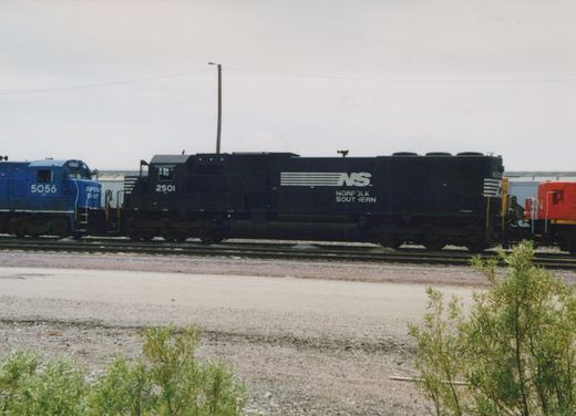 A black Norfolk Southern diesel locomotive numbered 2501 sits on tracks in a yard, with a blue locomotive and a red locomotive partially visible beside it, under a cloudy sky.