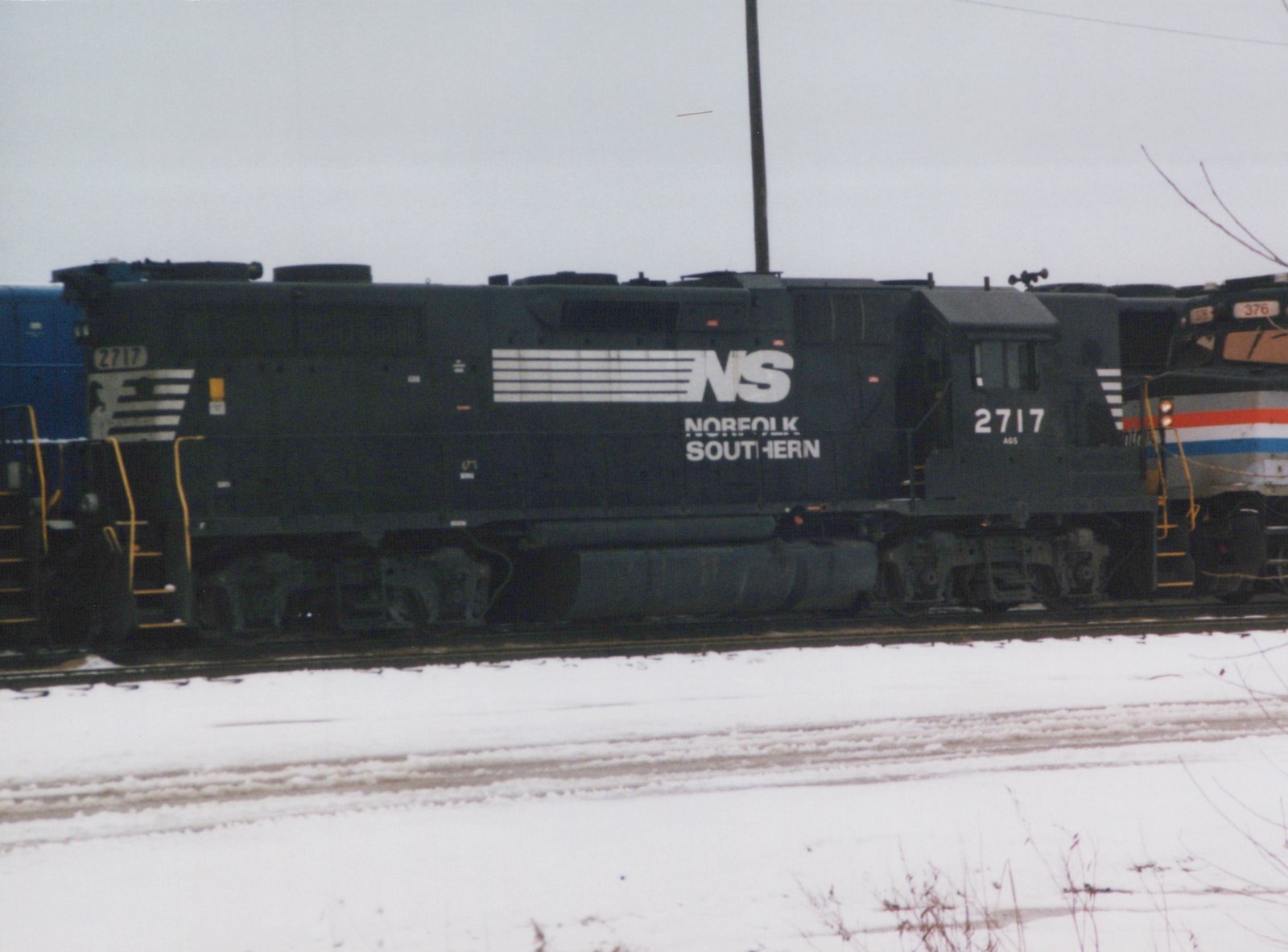 A black Norfolk Southern diesel locomotive, numbered 2717, sits on tracks in a snowy yard; part of another locomotive with a red, white, and blue livery is visible behind it.