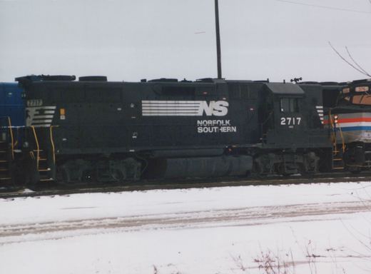 A black Norfolk Southern diesel locomotive, numbered 2717, sits on tracks in a snowy yard; part of another locomotive with a red, white, and blue livery is visible behind it.