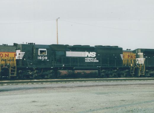 Side view of a black Norfolk Southern diesel locomotive numbered 1609, parked in a rail yard with a yellow locomotive behind it under a gray sky.