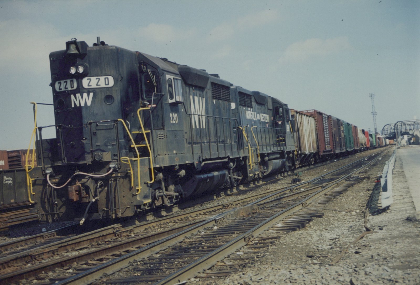 A black Norfolk and Western diesel locomotive numbered 220 is shown pulling a line of colorful freight cars on parallel tracks in a rail yard under a cloudy sky.