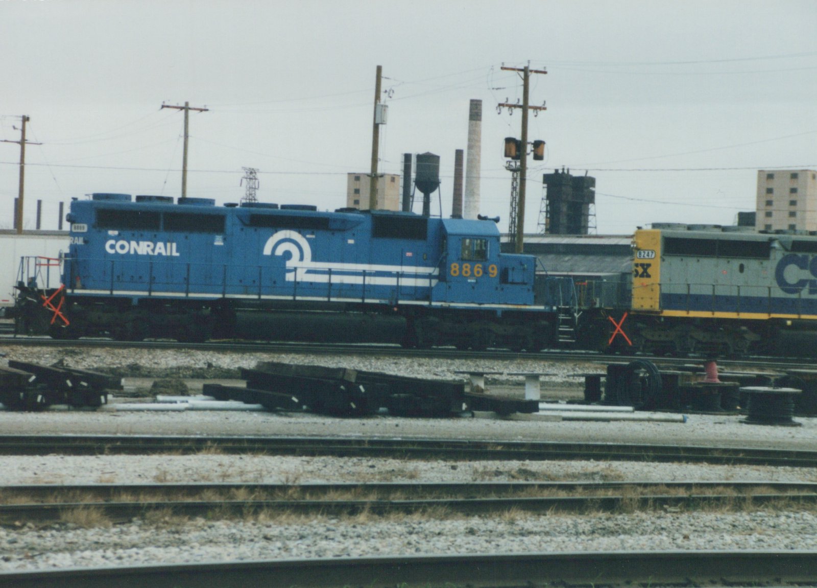 Side view of a blue Conrail locomotive with number 8869 parked next to a gray CSX locomotive with number 8247 in a gravel rail yard with industrial buildings in the background.