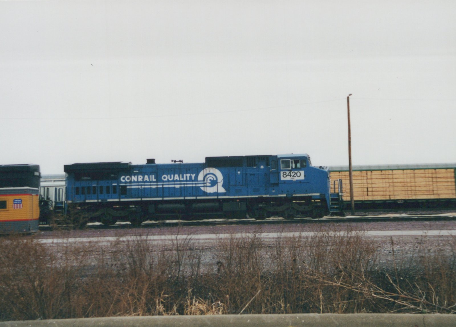 A side view of a blue Conrail diesel locomotive, number 8420, in a rail yard during winter, with freight cars behind it and bare brush in the foreground.
