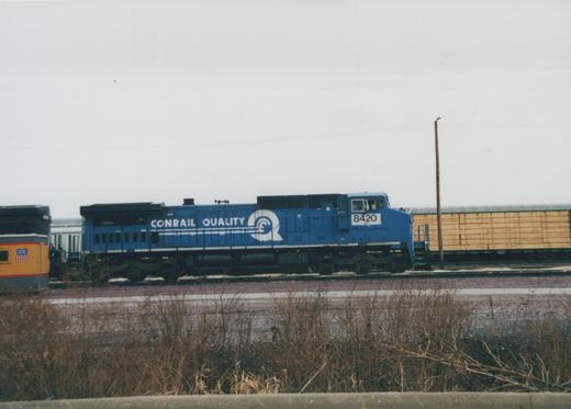 A side view of a blue Conrail diesel locomotive, number 8420, in a rail yard during winter, with freight cars behind it and bare brush in the foreground.