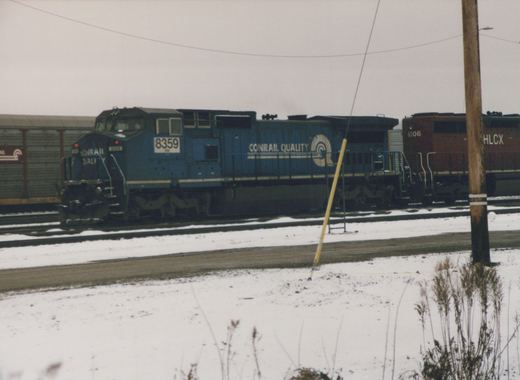 A blue Conrail locomotive with the number 8359 is parked on snowy tracks in a rail yard, with a brown HLCX locomotive numbered 6306 behind it.