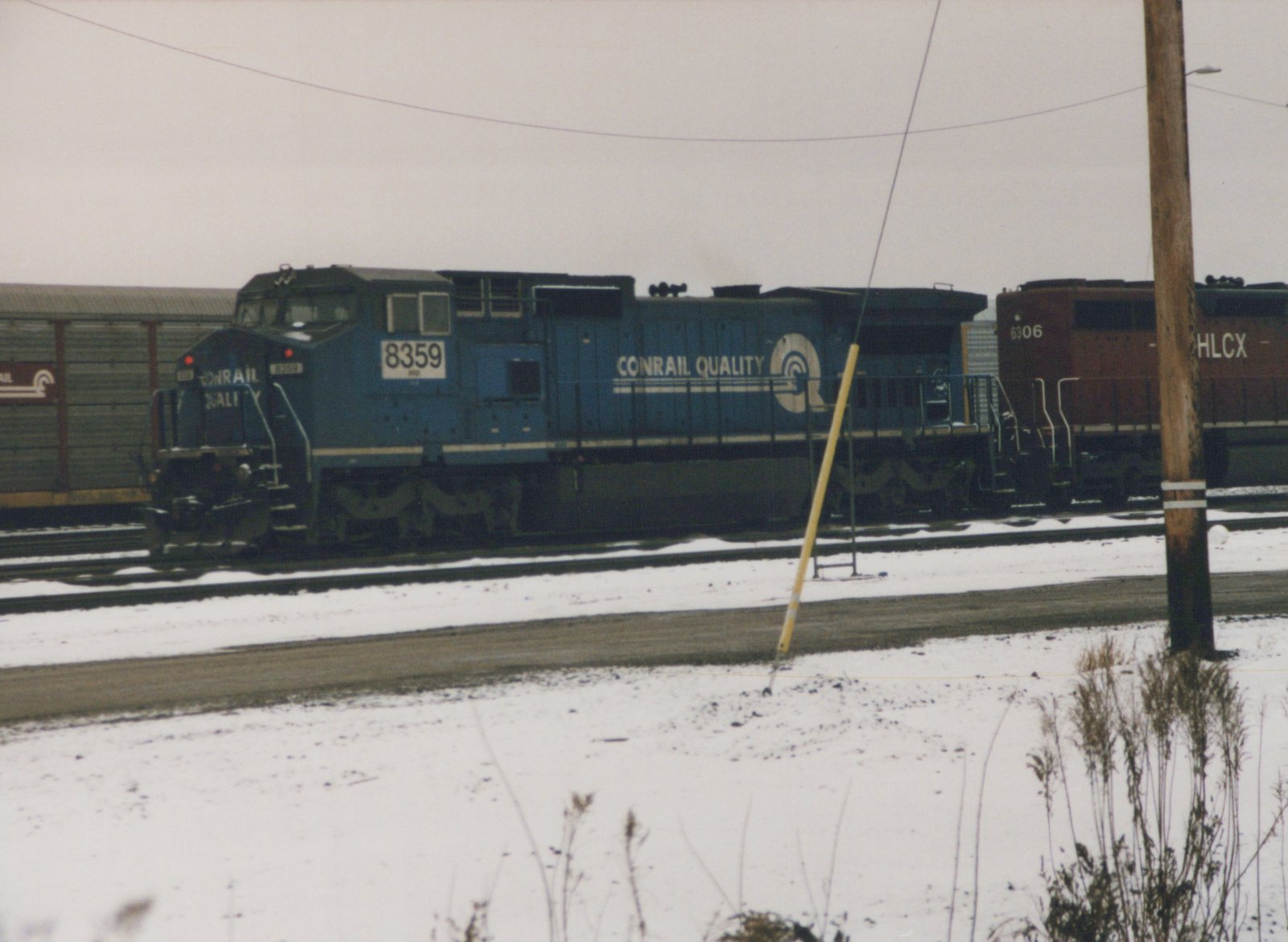A blue Conrail locomotive with the number 8359 is parked on snowy tracks in a rail yard, with a brown HLCX locomotive numbered 6306 behind it.