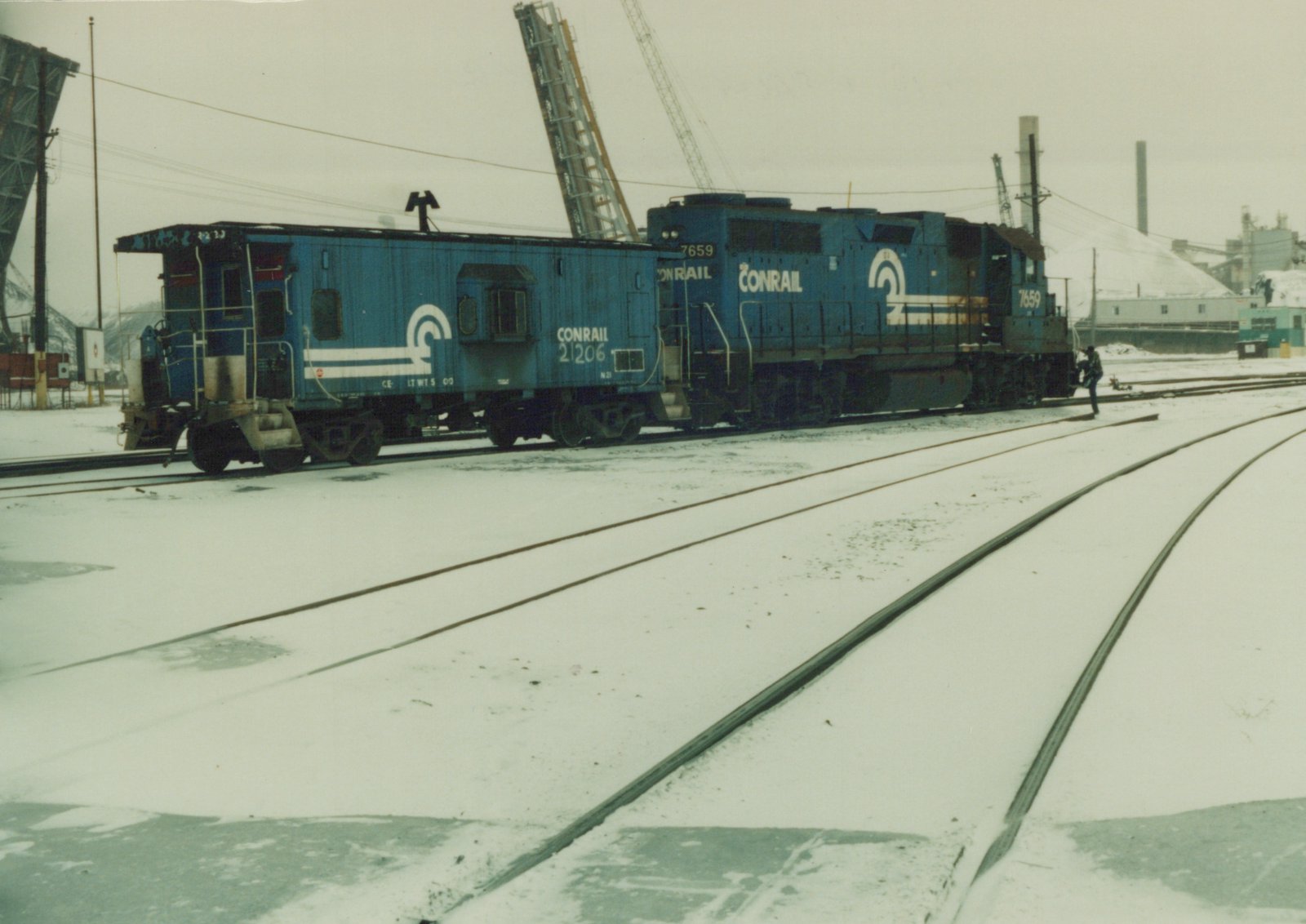A blue Conrail locomotive numbered 7659 pulls a matching caboose numbered 21206 through a snow-covered rail yard, with a single person standing near the locomotive.