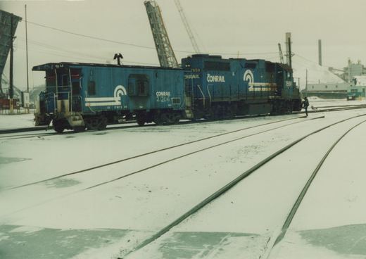 A blue Conrail locomotive numbered 7659 pulls a matching caboose numbered 21206 through a snow-covered rail yard, with a single person standing near the locomotive.
