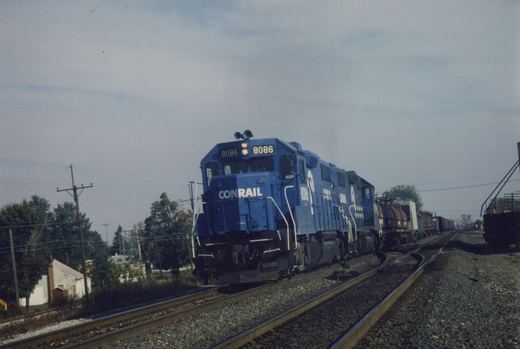 A blue Conrail diesel locomotive numbered 8086 is pulling a line of freight cars along a curving track, with trees showing autumn colors in the background.