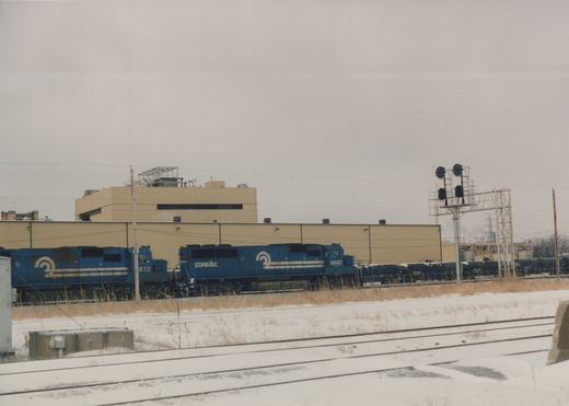 A blue Conrail locomotive with the number 6842 is pulling a line of freight cars along a snow-covered track with a large building in the background.
