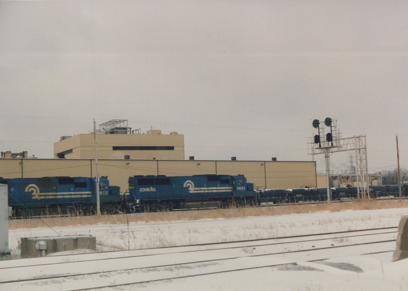 A blue Conrail locomotive with the number 6842 is pulling a line of freight cars along a snow-covered track with a large building in the background.
