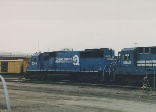 Side view of two blue Conrail diesel locomotives, numbered 6765 and 5060, standing on tracks in a rail yard under an overcast sky.