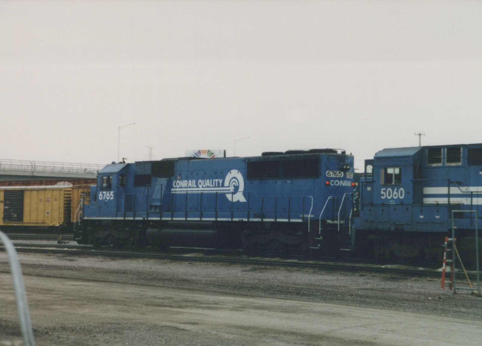 Side view of two blue Conrail diesel locomotives, numbered 6765 and 5060, standing on tracks in a rail yard under an overcast sky.