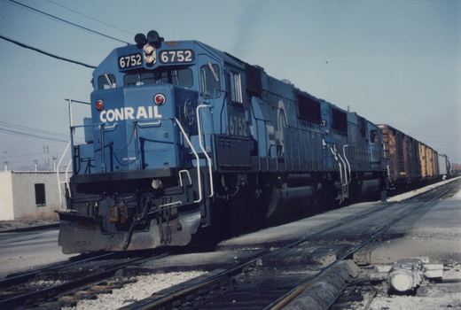 A blue Conrail GP38-2 diesel locomotive, number 6752, is parked on a set of tracks in a yard, with a line of freight cars behind it.
