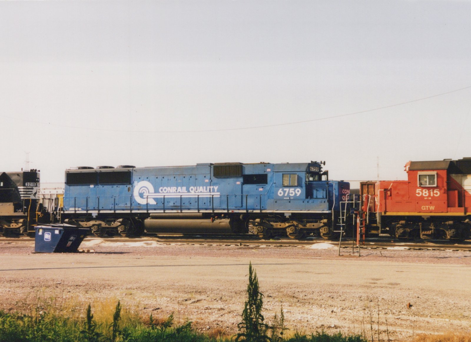 A blue Conrail locomotive with the number 6759 is parked on tracks in a rail yard, with a red GTW locomotive and a black locomotive visible nearby.