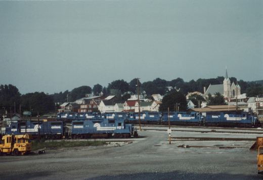 Several blue Conrail locomotives are parked side-by-side on multiple tracks in a gravel yard, with residential houses and a church steeple visible in the tree-lined background under a gray sky.