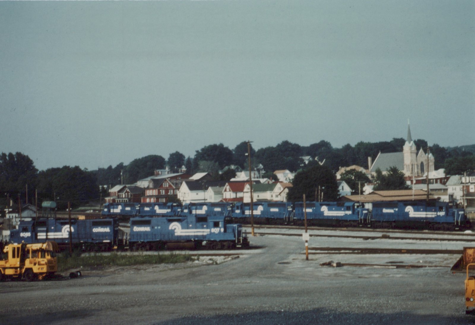 Several blue Conrail locomotives are parked side-by-side on multiple tracks in a gravel yard, with residential houses and a church steeple visible in the tree-lined background under a gray sky.