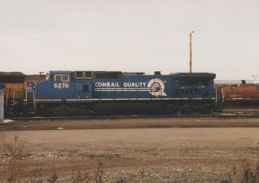 A blue Conrail diesel locomotive, number 6276, sits on the tracks in a gravel yard under an overcast sky.