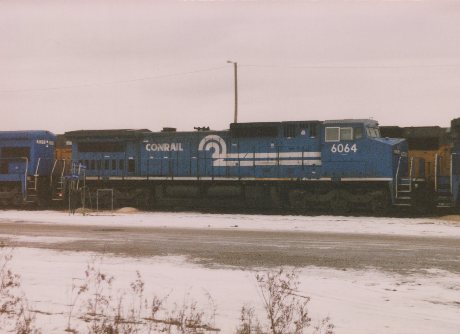 A side view of a blue Conrail diesel locomotive, number 6064, parked on a snowy railyard with another Conrail locomotive partially visible beside it.