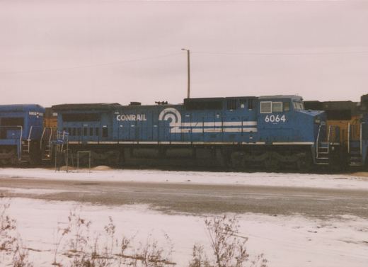 A side view of a blue Conrail diesel locomotive, number 6064, parked on a snowy railyard with another Conrail locomotive partially visible beside it.
